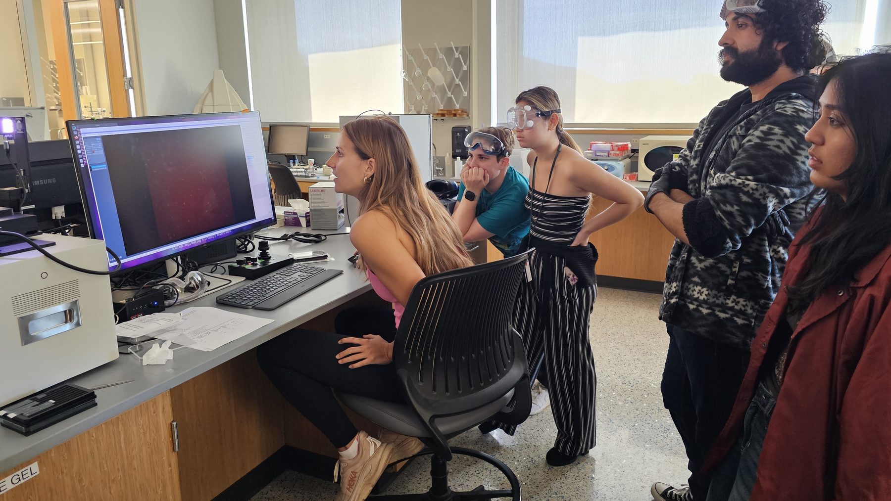 A group of students and lab members watching a microscope image on a computer screen while one student sits at the controls