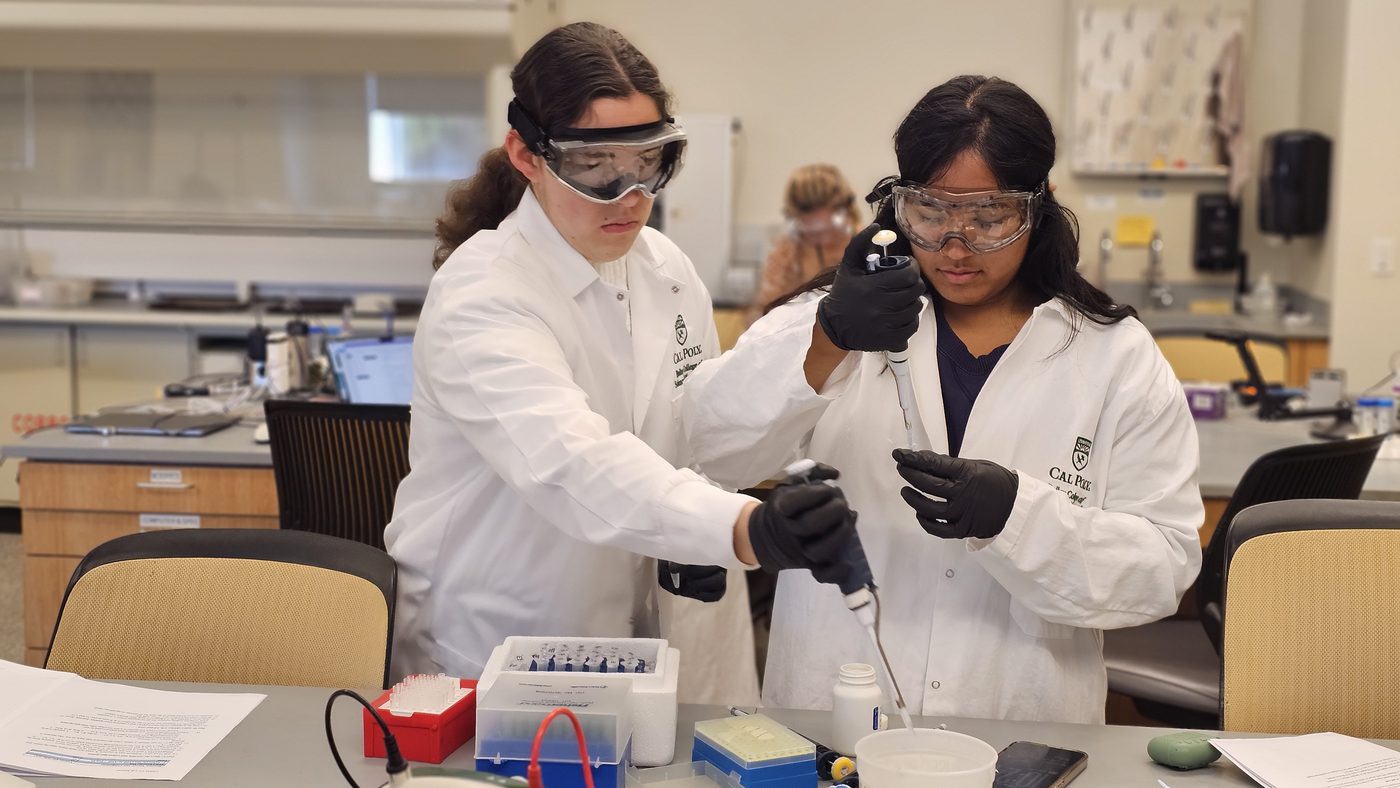 Two students in lab coats, safety goggles and gloves pipetting together at a lab bench