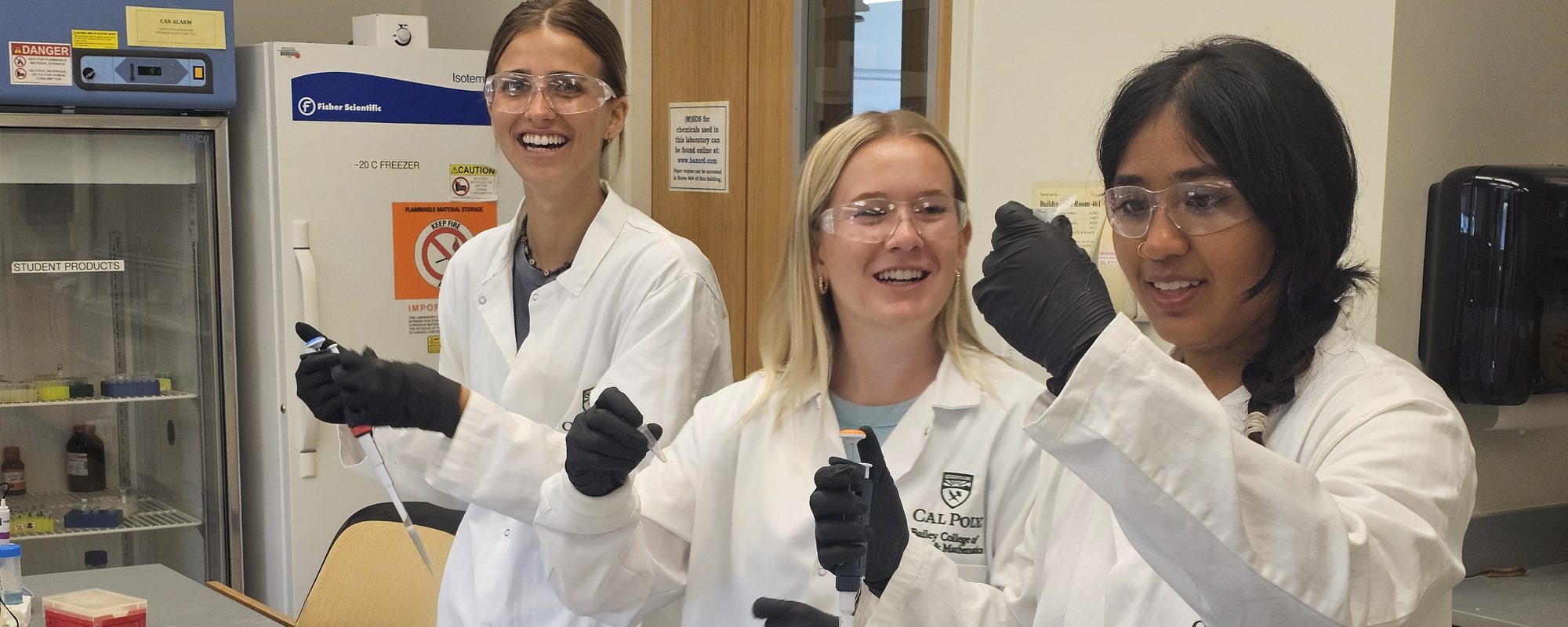 Three students in lab coats, safety glasses and gloves laughing as they pipette at a laboratory bench