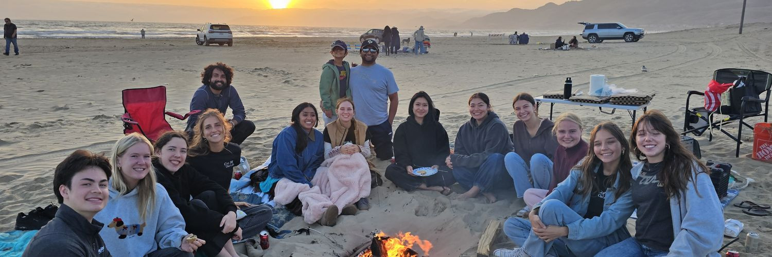 Members of the Oza Lab, including Javin Oza standing at the back, gathered around a beach bonfire at sunset and smiling at the camera