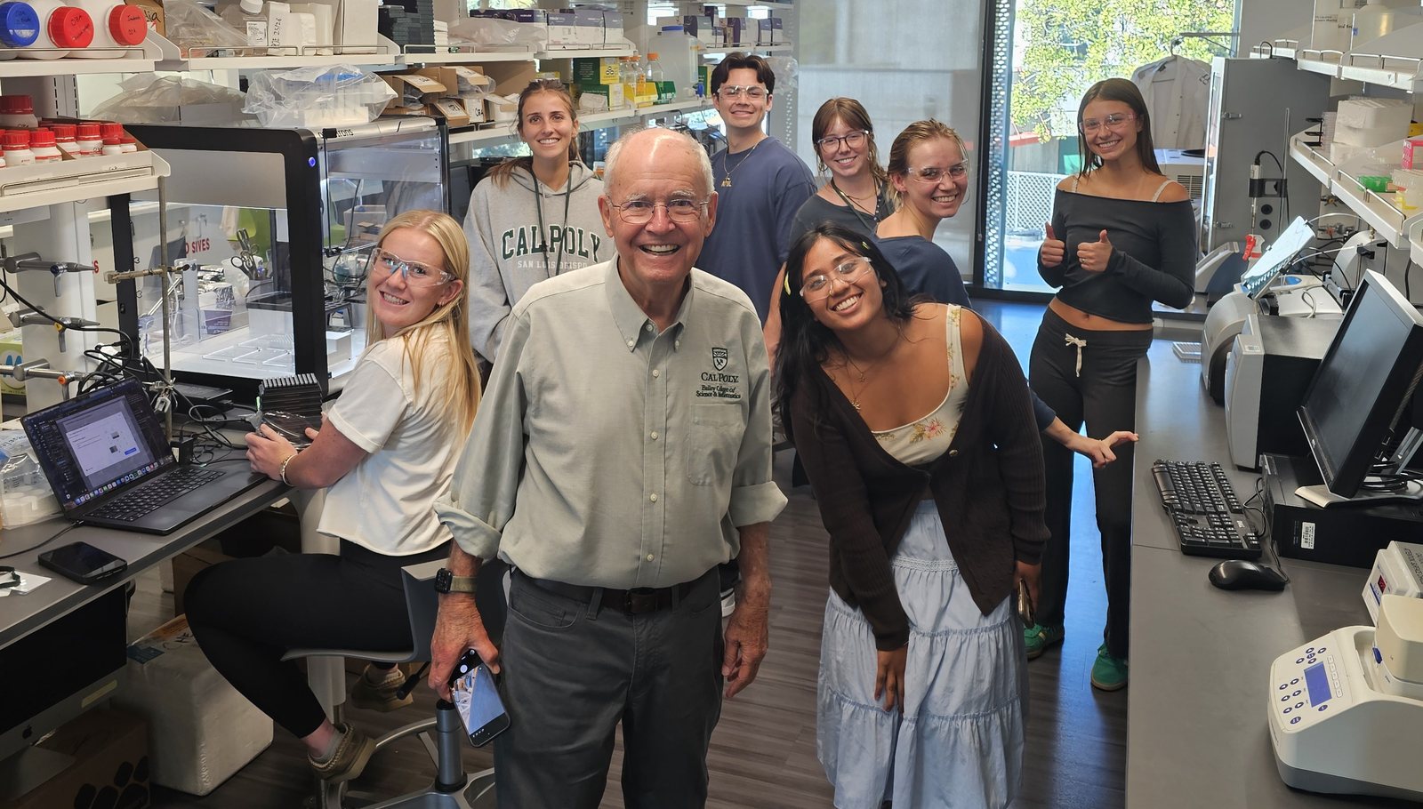 Members of the Oza Lab smiling for a photo among shelves of reagents and benchtop equipment, wearing safety glasses, one giving two thumbs up
